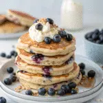 Fluffy blueberry pancake protein stack topped with whipped cream and fresh blueberries on a white plate.