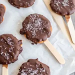 Homemade high-protein Dilly Bars coated in rich chocolate and sprinkled with sea salt on parchment paper.