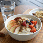 Overnight oats with water topped with fresh fruit, granola, and cinnamon in a white bowl beside a glass of water.