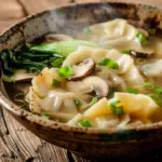 Steaming bowl of potsticker soup with mushrooms, bok choy, and scallions on a wooden table.